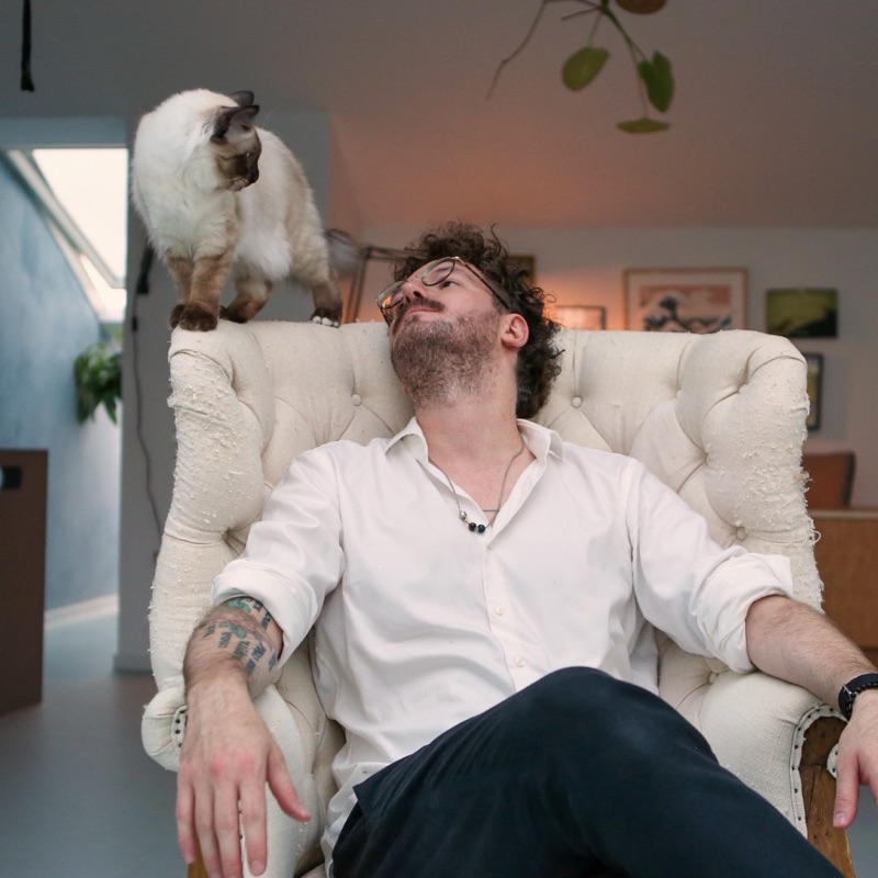 Patrick im Sessel, auf der Rückenlehne steht eine helle Katze mit dunklen Ohren und Pfoten